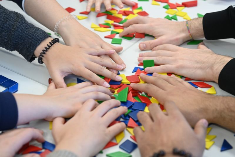 Mehrere Hände strecken sich aus, um bunte geometrische Plastikformen zu berühren und aufzuheben, die auf einem weißen Tisch verstreut sind.
