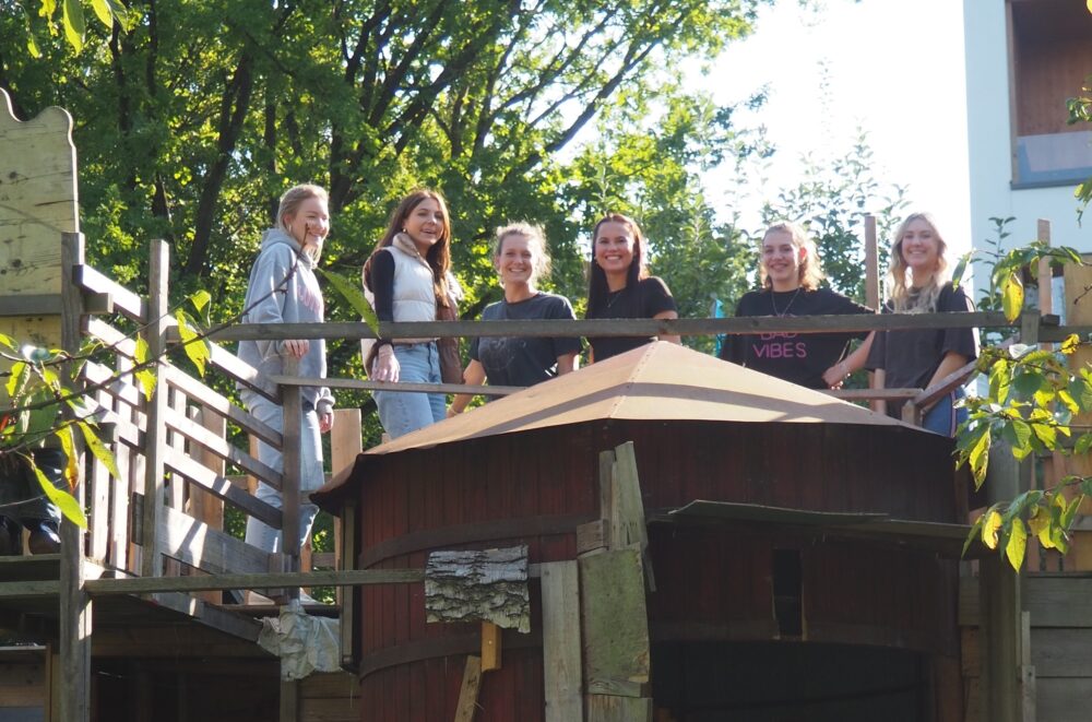 Sechs lächelnde junge Frauen stehen gemeinsam auf einem Holzspielgerät auf dem Bauspielplatz Regensburg, umgeben von Bäumen und Sonnenlicht, und posieren für das Foto.