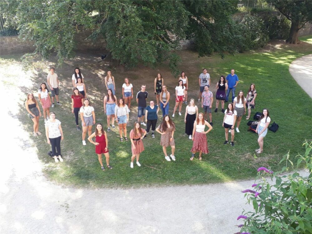 Eine Gruppe junger Erwachsener steht draußen auf einer Wiese, mit Abstand zueinander und blickt in die Kamera. Im Hintergrund sind Bäume und ein Weg zu sehen. Die Formation ähnelt einem Herzen.