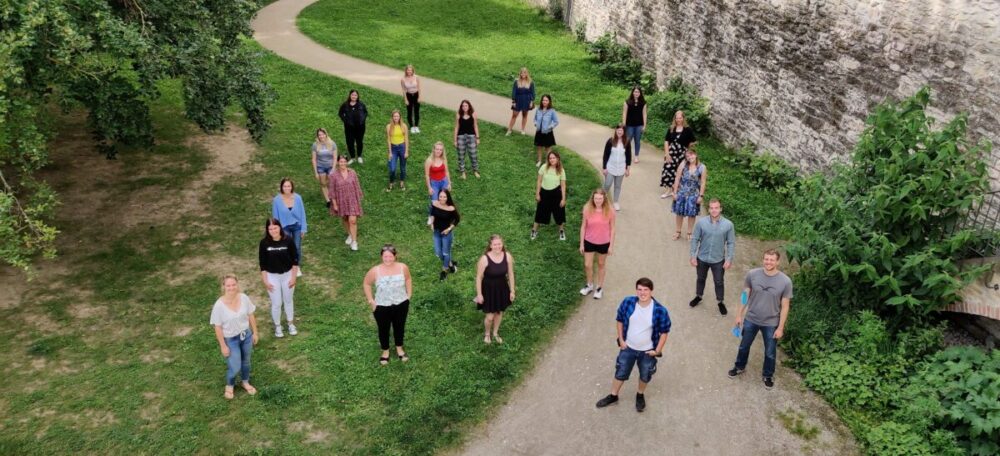 Eine Gruppe von Menschen steht mit Abstand auf Gras und einem Gehweg in der Nähe einer Steinmauer und bildet die Form eines Herzens. Im Hintergrund sind Bäume und Grün zu sehen. Das Foto wurde von oben aufgenommen.