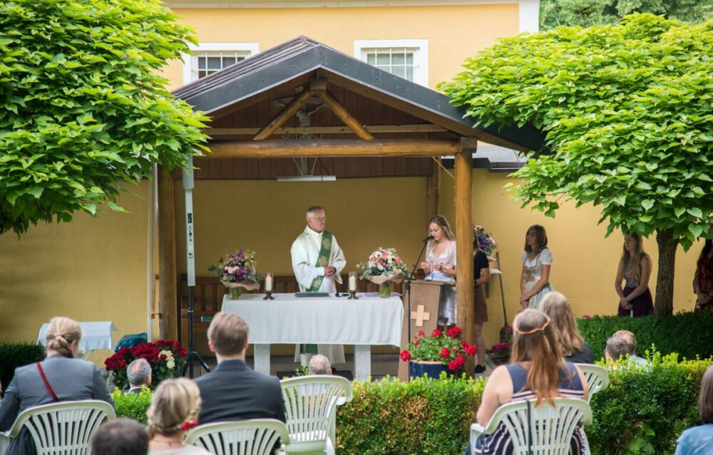 Eine Hochzeitszeremonie im Freien mit einem Priester am Altar und einer Frau, die am Podium spricht. Die Gäste sitzen auf weißen Stühlen mit Blick auf den Altar, umgeben von Grün und Blumen. Im Hintergrund ist ein gelbes Gebäude zu sehen.