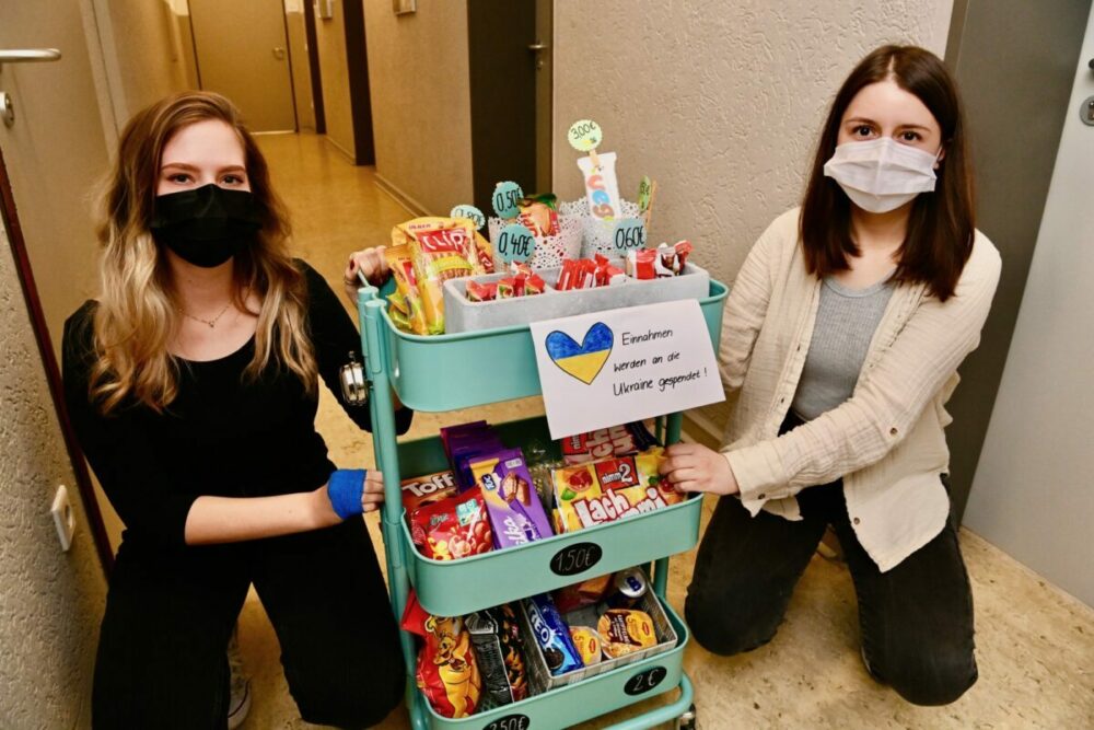 Zwei junge Frauen mit Masken knien neben einem blaugrünen Wagen voller Snacks und Getränke. Auf einem Schild mit einem blau-gelben Herz steht auf Deutsch: „Einsamem kann so oft die Ukraine geholfen!“. Die Snacks sind mit Preisschildern versehen.
