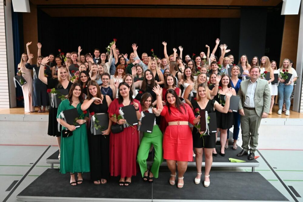 Eine große Gruppe von Menschen, hauptsächlich Frauen in Abendgarderobe, steht auf gestuften Podesten im Innenbereich. Viele halten Rosen in den Händen, winken und lächeln in die Kamera, was auf eine Feier oder eine Abschlussfeier schließen lässt.