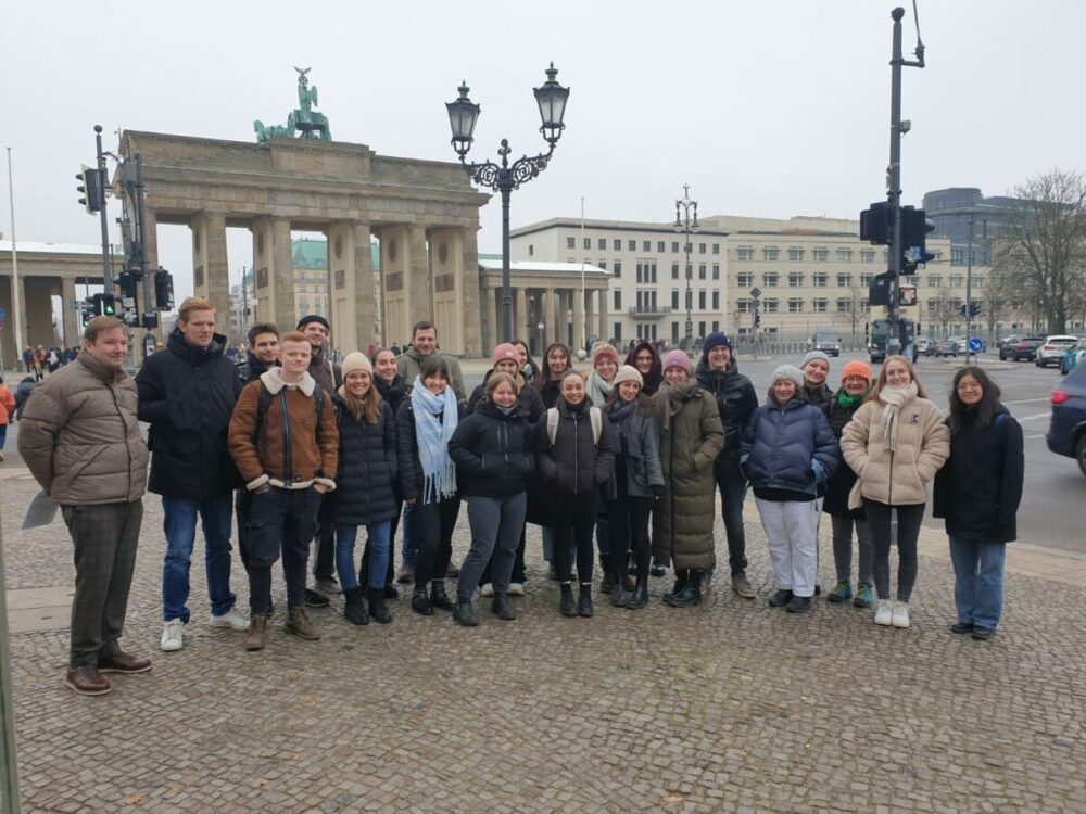 Eine Gruppe von Menschen in Winterkleidung posiert für ein Foto vor dem Brandenburger Tor in Berlin. Der Himmel ist bedeckt, im Hintergrund sind Gebäude und Straßenlaternen zu sehen.