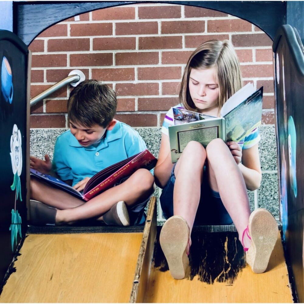 Zwei Kinder, ein Junge und ein Mädchen, sitzen nebeneinander auf einer Spielplatzrutsche und lesen konzentriert Bücher. Im Hintergrund ist eine Backsteinmauer zu sehen. Beide scheinen sich auf ihre Bücher zu konzentrieren und haben die Beine ausgestreckt.
