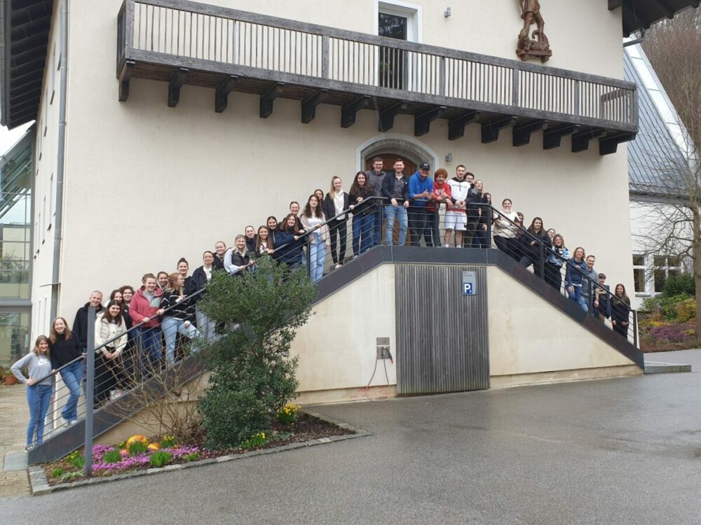 Eine große Gruppe von Menschen steht auf beiden Seiten einer Außentreppe, die an ein beiges Gebäude angrenzt, und lächelt in die Kamera. Im Vordergrund sind einige Pflanzen und bunte Blumen zu sehen.