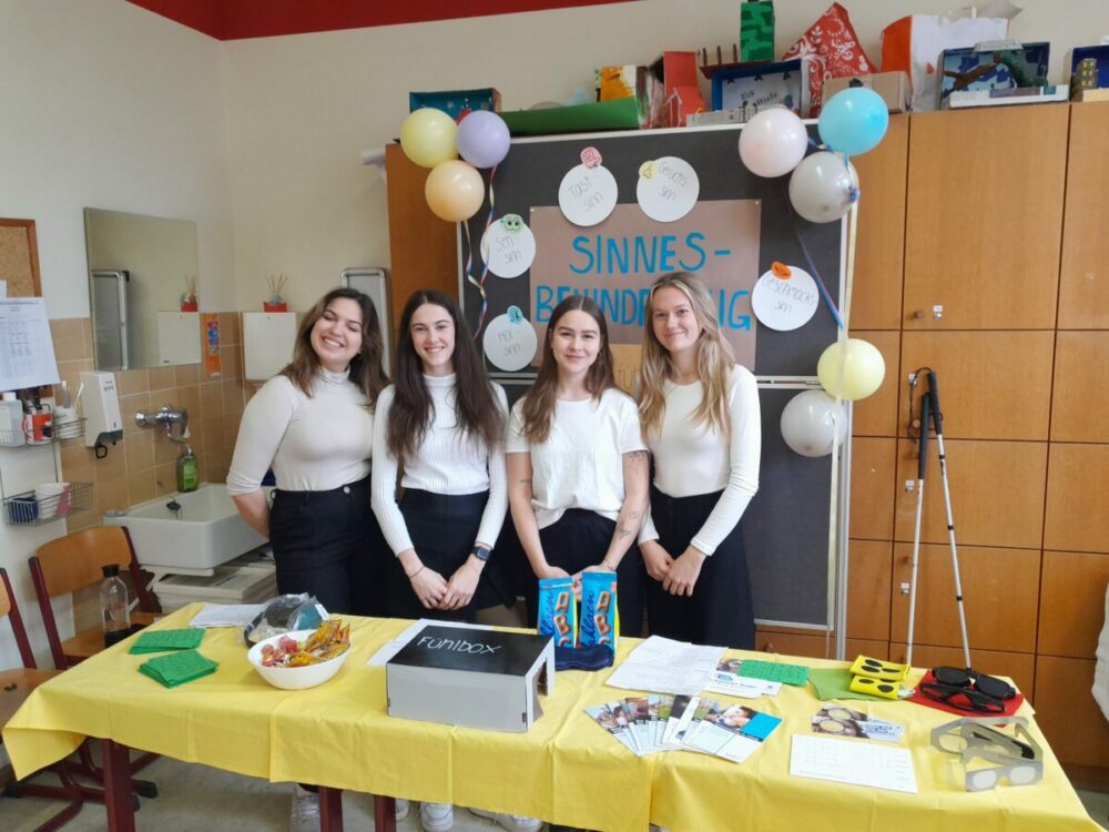 Vier junge Frauen stehen hinter einem dekorierten Tisch mit sensorischen Aktivitätsmaterialien und lächeln in die Kamera. Hinter ihnen hängt ein blaues Plakat mit Luftballons mit der Aufschrift „SINNES-BEWUNDERUNG“, und der Tisch ist mit bunten Gegenständen und Flyern bedeckt.