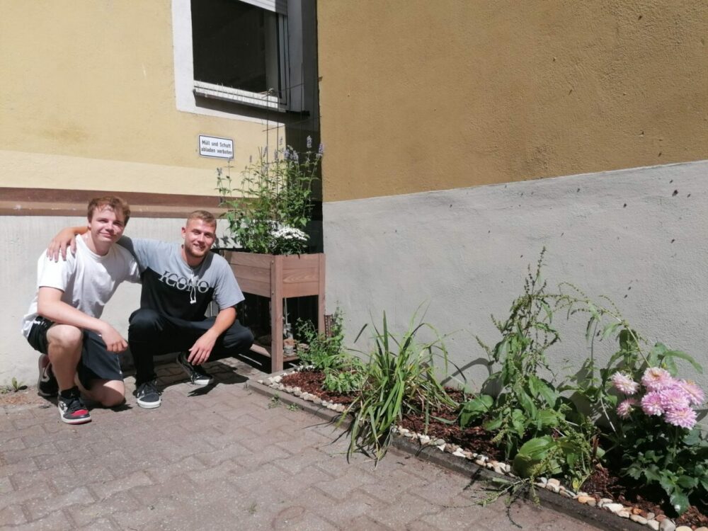 Zwei junge Männer hocken lächelnd neben einem kleinen Stadtgarten mit Blumen und Grünpflanzen vor einer gelb-grauen Hauswand. Sonnenlicht wirft Schatten, und an der Wand über ihnen ist ein kleines Schild zu sehen.