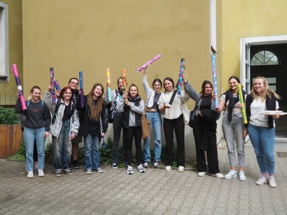 Eine Gruppe von zwölf jungen Leuten steht lächelnd vor einem gelben Gebäude und hält bunte Röhren in den Händen. Die meisten tragen Freizeitkleidung, die Stimmung ist fröhlich und feierlich.
