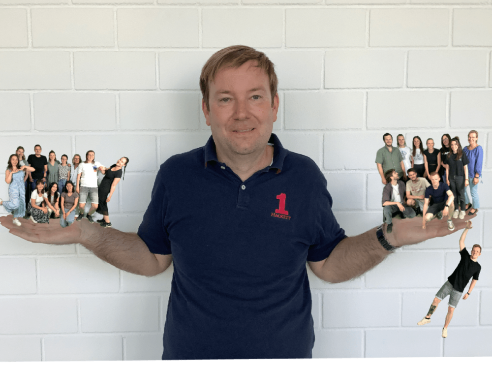 Ein Mann steht lächelnd mit ausgestreckten Armen vor einer weißen Backsteinmauer. Jede Hand scheint eine kleine Gruppe von Menschen zu halten, die auf seinen Handflächen angeordnet sind. Eine weitere kleine Person befindet sich in der Nähe seines rechten Ellbogens.