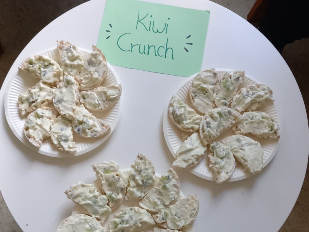 Drei Pappteller mit halbrunden Snacks, garniert mit einem cremigen weißen Aufstrich und Kiwistücken, stehen auf einem runden weißen Tisch. Ein grünes Schild mit der Aufschrift „Kiwi Crunch“ heißt die Kinder herzlich willkommen und bietet ihnen einen leckeren Snack für den Schulstart der Caritas Regensburg.