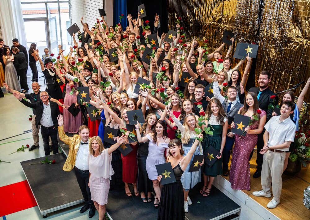Eine große Gruppe von Absolventen der Caritas Fachakademie Regensburg in Abendgarderobe feiert drinnen, hält Diplome und rote Rosen hoch, ist von goldenen Dekorationen umgeben und lächelt bei ihrer Abschlussfeier 2024 in die Kamera.