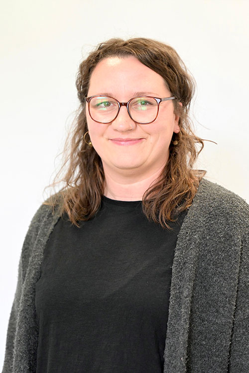 Eine Person mit gewelltem braunem Haar, die eine runde Brille, ein schwarzes Hemd und eine graue Strickjacke trägt, steht vor einem schlichten hellen Hintergrund und lächelt leicht in die Kamera.