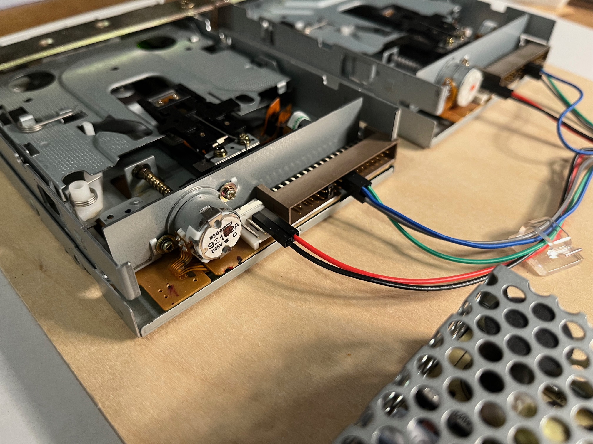 Nahaufnahme von zwei alten Diskettenlaufwerken, die über farbige Drähte mit einem Netzteil auf einer Holzfläche verbunden sind, mit elektronischen Bauteilen und Datenanschlüssen.