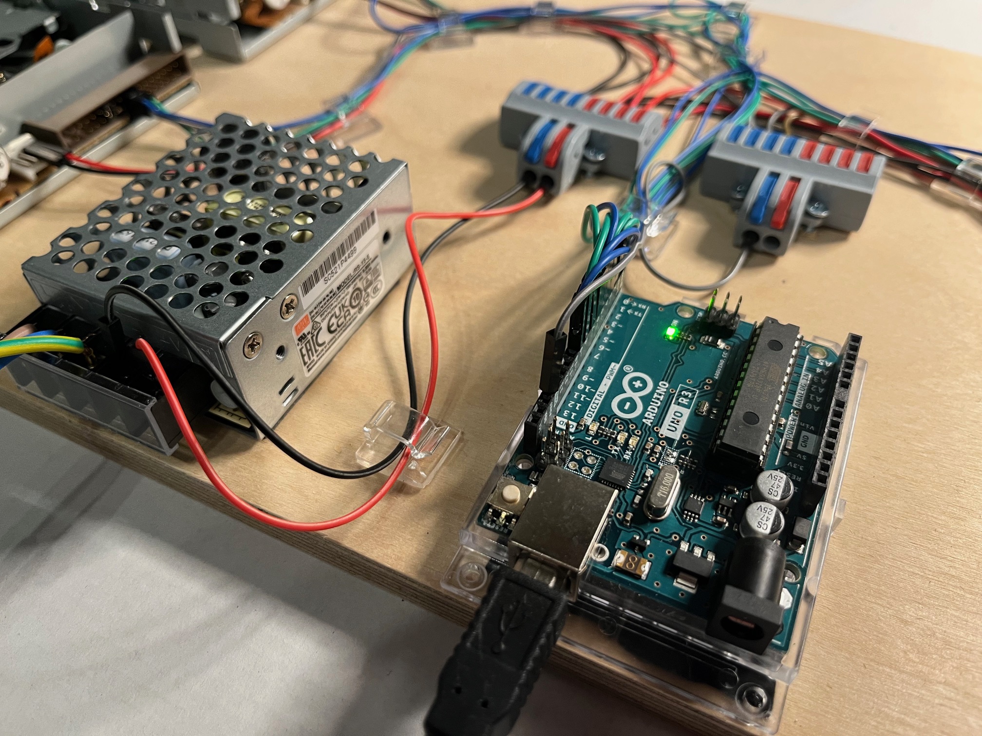Eine Nahaufnahme einer Arduino-Platine, die an ein Netzteil und zwei Klemmenleisten mit verschiedenfarbigen Drähten angeschlossen ist, die alle auf einem Holzbrett montiert sind. Die grüne LED des Arduinos leuchtet und zeigt damit an, dass er eingeschaltet ist.