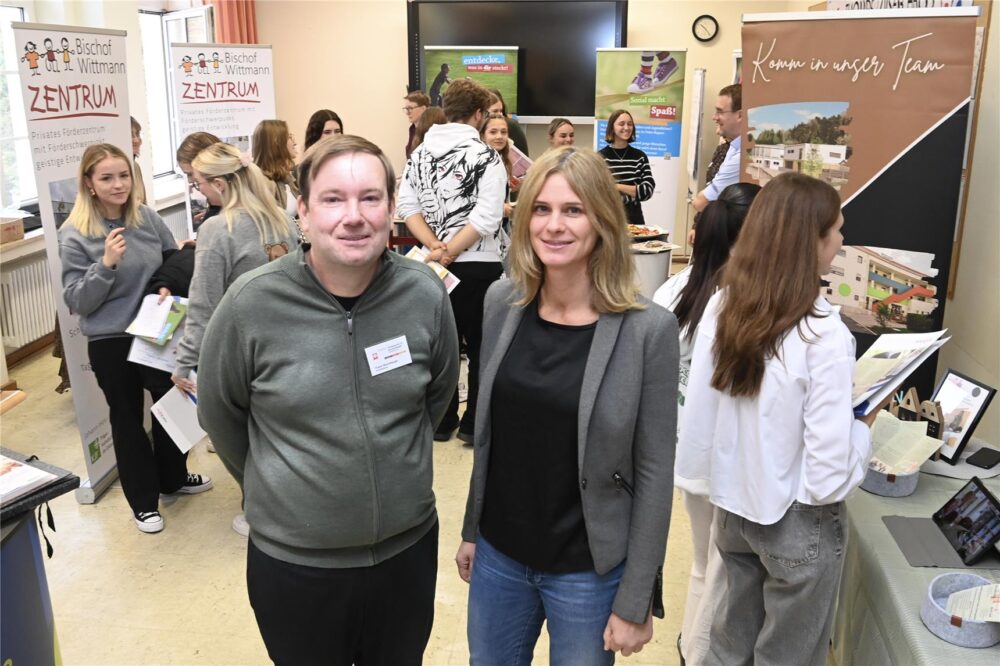 Zwei lächelnde Personen stehen auf einer gut besuchten Hausmesse der Caritas FAKS im Oktober mit Tischen, Postern und Broschüren. Andere Teilnehmer interagieren im Hintergrund mit Arbeitgebern und erkunden Möglichkeiten für Erzieher.