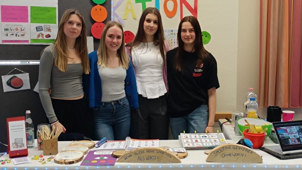 Vier junge Frauen stehen hinter einem bunten Stand auf dem PPH Heilpädagogik Marktplatz. Auf dem Tisch liegen Schilder, Flyer und Bastelmaterialien. Sie lächeln für das Foto, umgeben von Klassenzimmerplakaten und Dekorationen im Hintergrund.