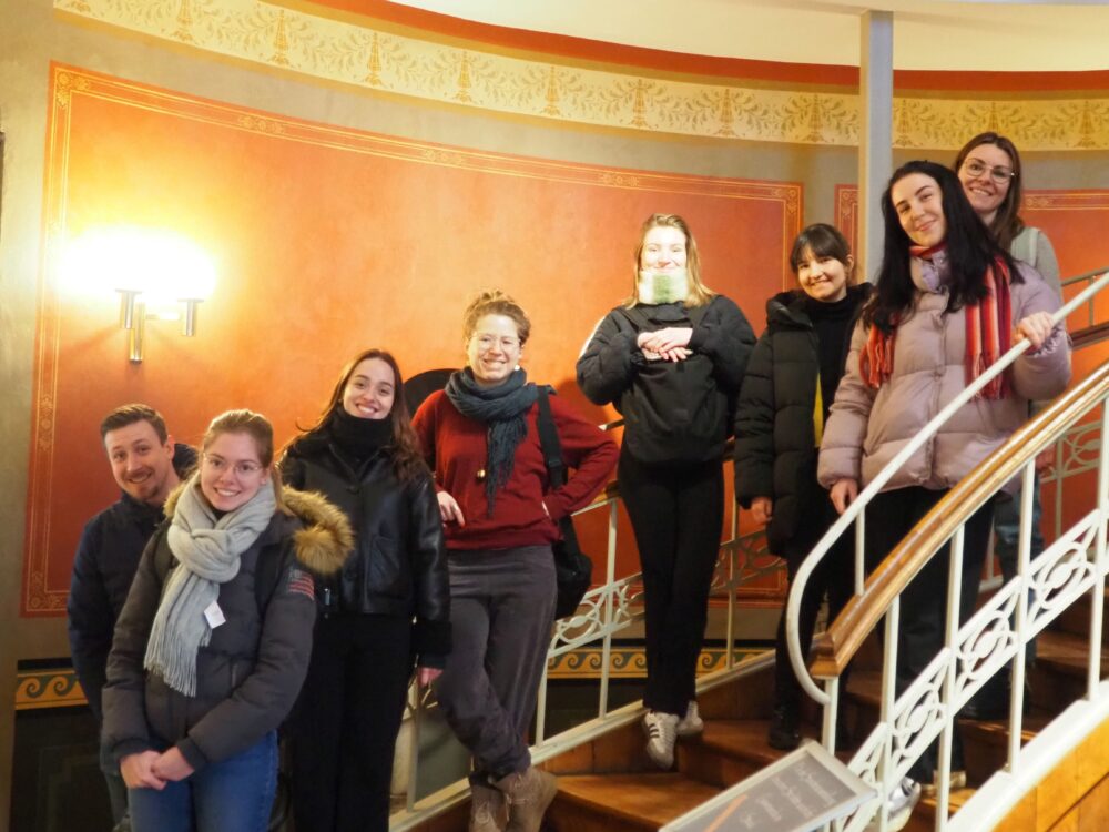 Eine Gruppe von acht in Winterkleidung eingemummelten Personen steht lächelnd auf einer Wendeltreppe mit verziertem Geländer im warm beleuchteten, reich dekorierten Innenraum eines Naturkundemuseums.