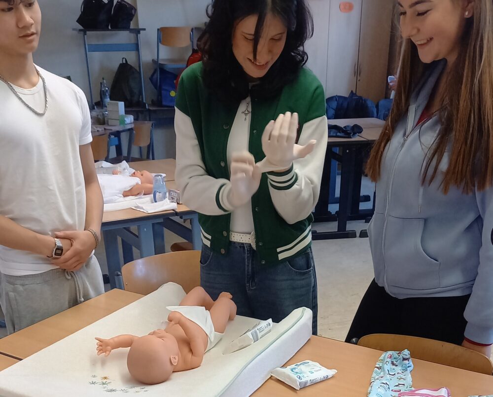 Drei Jugendliche stehen mit einer Babypuppe, Windeln und Feuchttüchern um einen Tisch. Eine Person zieht Handschuhe an, um das Wickeln im Rahmen der SEJ Säuglingspädagogik zu demonstrieren, während die anderen lächelnd im Klassenzimmer zuschauen.