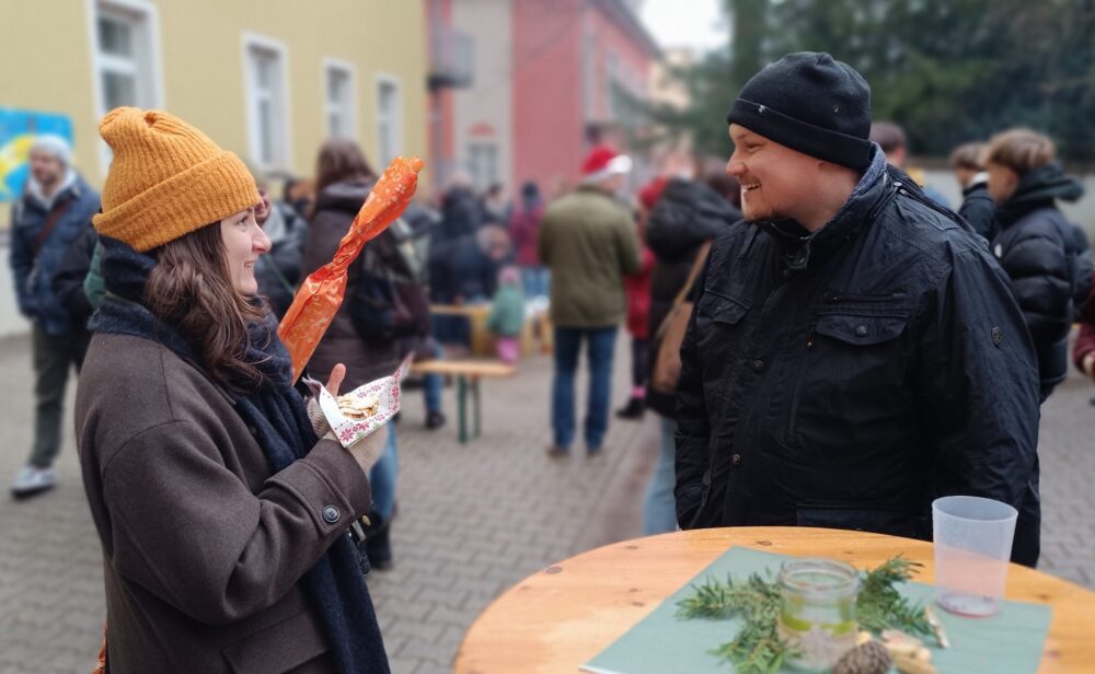 Bei einer lebhaften Caritas-Veranstaltung im Innenhof überreicht eine Frau mit gelbem Hut einem lächelnden Mann in dunkler Kleidung ein verpacktes Geschenk, im Hintergrund sind Menschen und Tische vom Weihnachtsmarkt zu sehen.